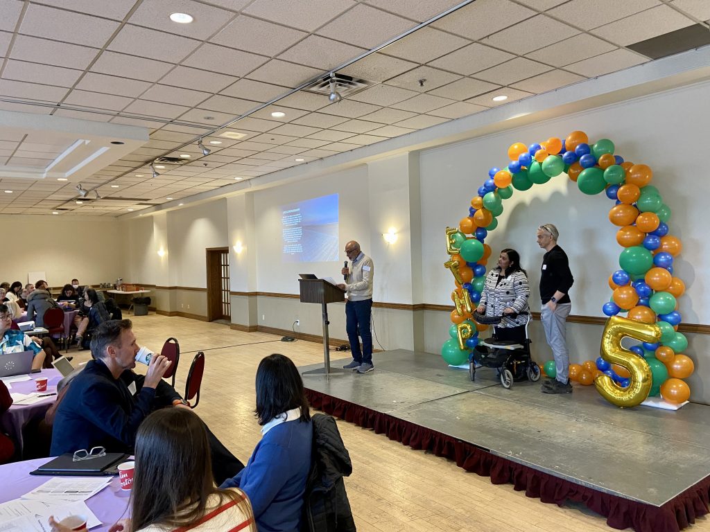 Three people on a small stage speaking while an audience looks on.