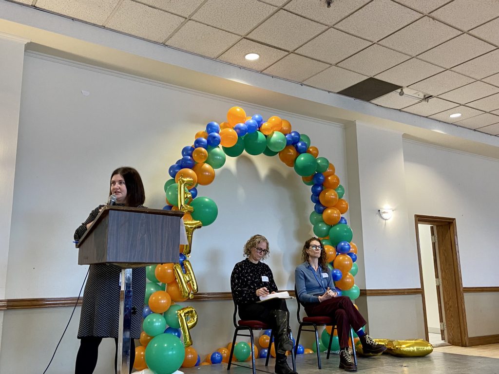 A person speaking at a podium while two other people sit nearby under a balloon arch.